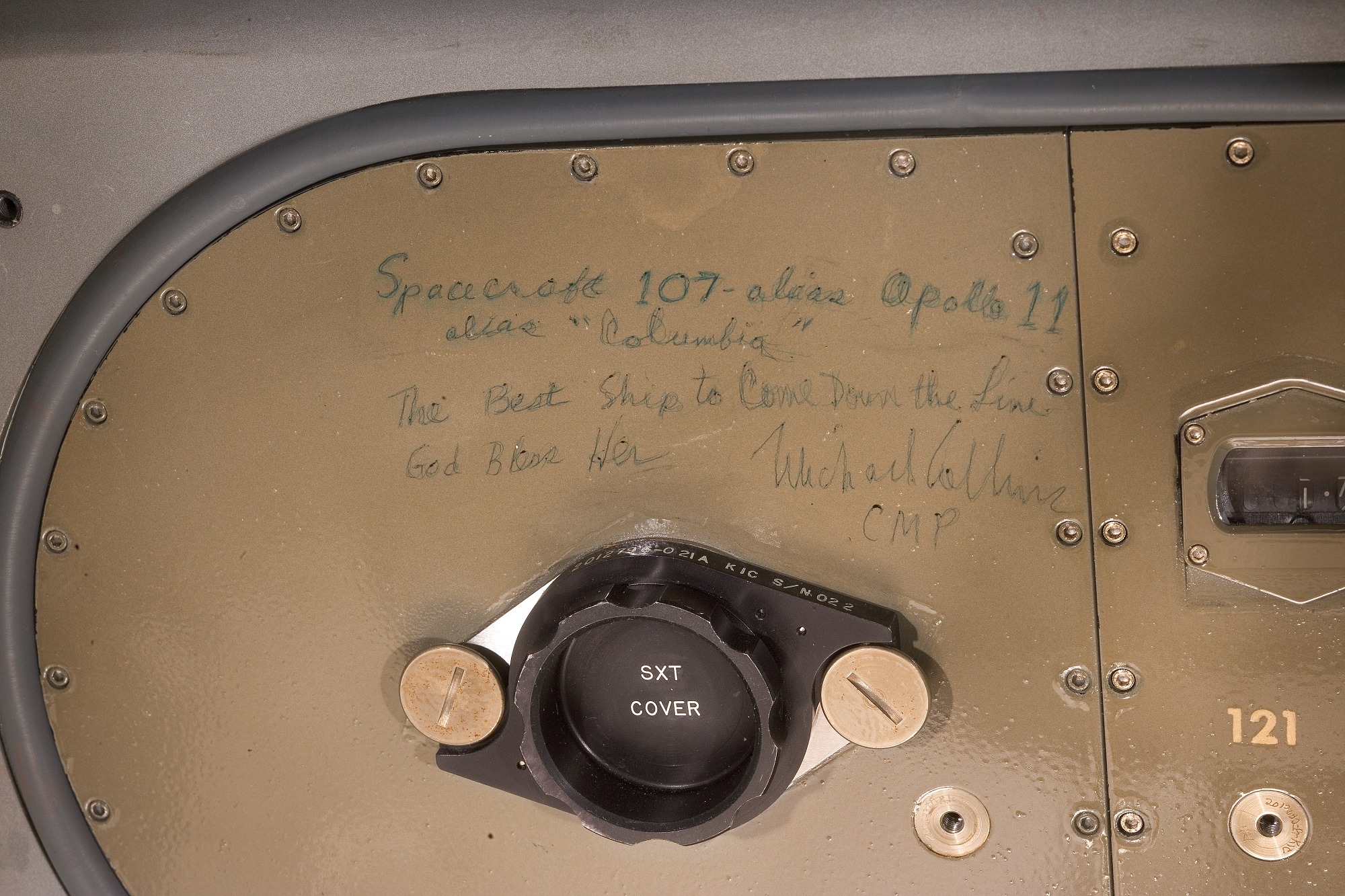 Metal panel with rivets around the edge. Handwritten text in green ink reads: ‘Spacecraft 107 – alias Apollo 11 / alias “Columbia” / The Best Ship to Come Down the Line / God Bless Her / Michael Collins / CMP’.